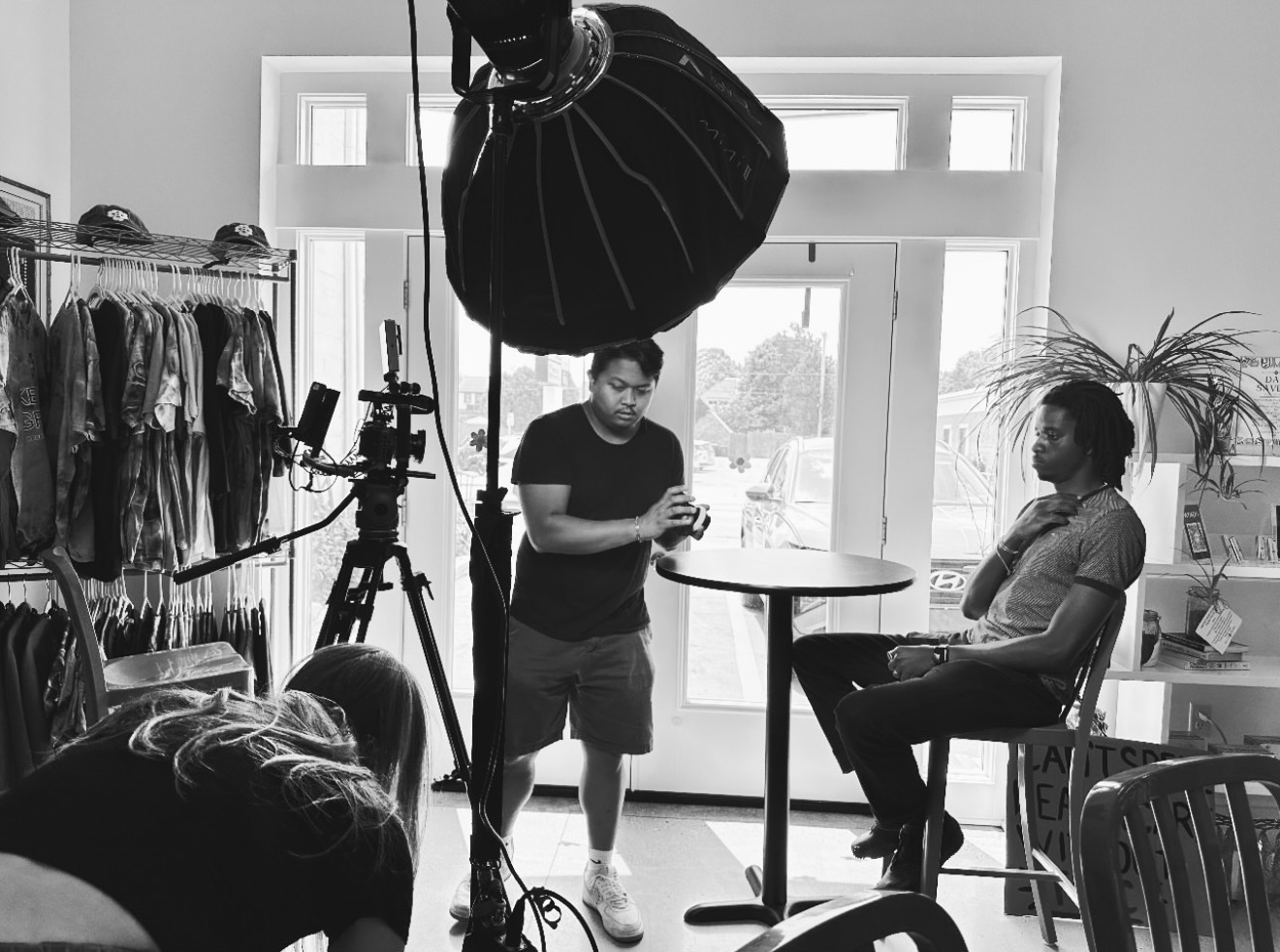 Nova Hill crew filming an on-camera interview with a large softbox light, cinema camera on tripod, camera operator, and seated subject inside a retail location.